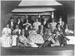 Cast of a dramatic production with men dressed as ladies; some holding fans.